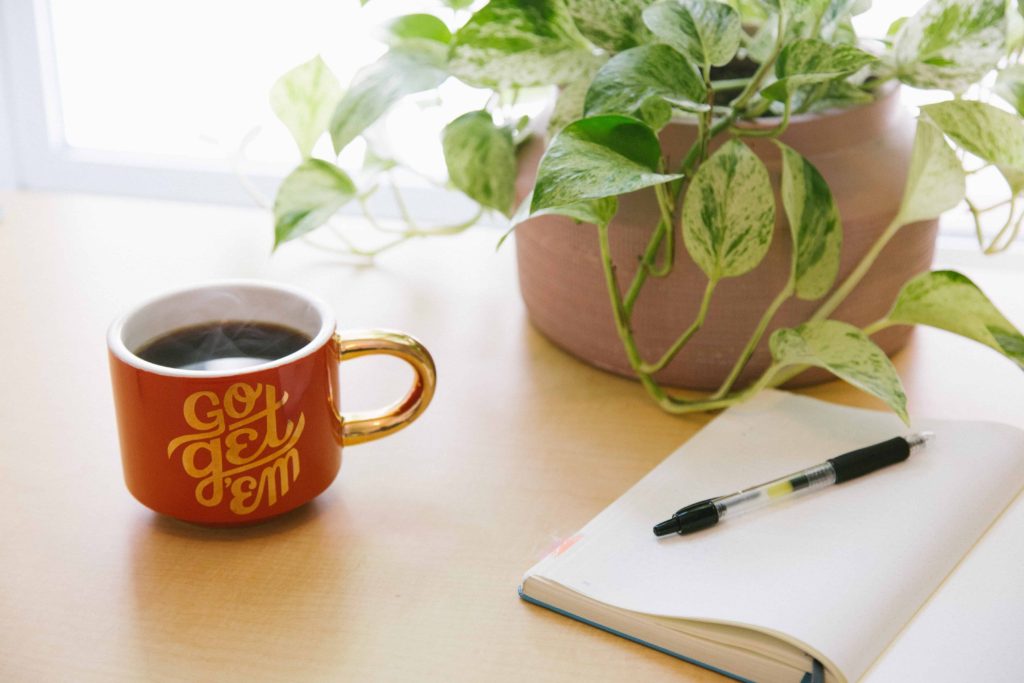 red coffee mug that says go get em on desk by plant and notebook with pen