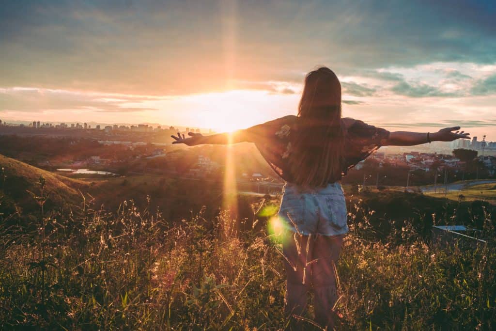 girl on grassy open hill watching sunset over city skyline with outstretched arms