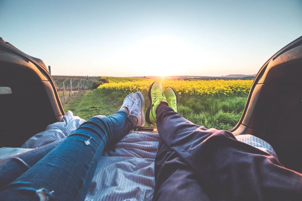 Two people laying in the back of the the car watching the sunset over a yellow field