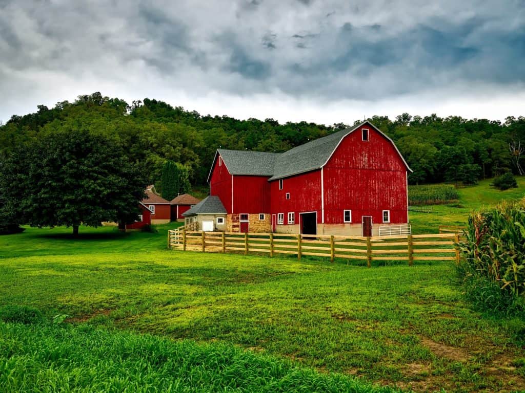 Red picturesque barn in green field with fences near woods and hill