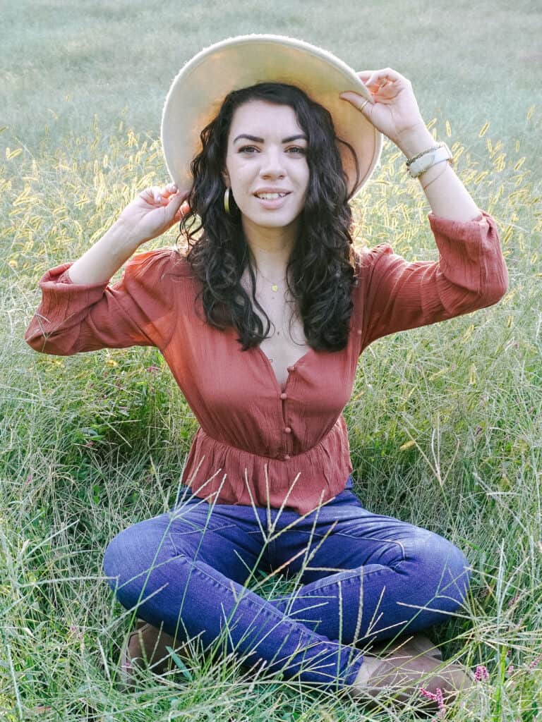 brunette holding a farm hat while sitting in a grassy field